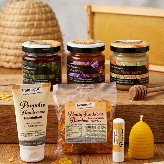 Ein Blick auf das "Kleine Kennlernpaket" der Imkerei Ostermann, präsentiert auf einem rustikalen Holzbrett vor einem Bienenkorb. Es zeigt drei 50g Gläser regionalen Honigs (Waldhonig, Sommerhonig, Hugsweierer Blütenzauber), einen Beutel Honig-Sanddorn-Bärchen, eine Tube Propolis-Handcreme, einen Propolino-Lippenpflege-Stift und eine Bienenwachskerze in Bienenkorb-Form. 