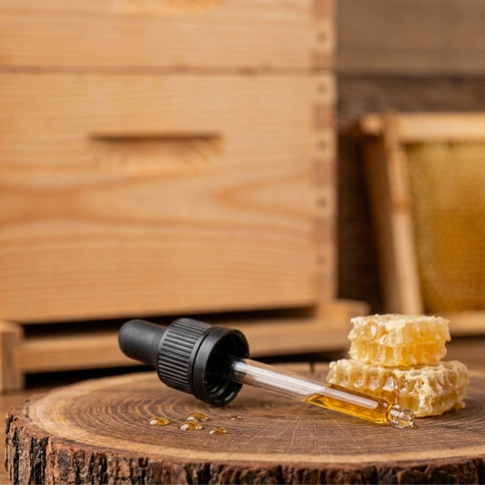 Hochwertige Glaspipette mit schwarzem Saugkopf auf einer rustikalen Holzscheibe neben frischen Bienenwaben vor einem Bienenstock-Hintergrund.
