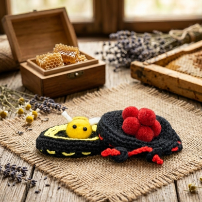 Zwei handgefertigten Häkel-Deckelhauben auf Sackleinen. Links: Ein Häkel-Häubchen in Bienenform. Rechts: Ein Häkel-Häubchen im Schwarzwälder Bollenhut-Stil mit roten Wollbollen. Im Hintergrund eine Holzkiste mit Honigwaben.