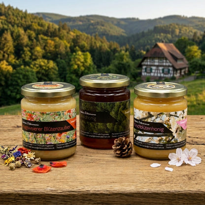 Drei Gläser Honig (Hugsweierer Blütenzauber, Waldhonig, Kirschblütenhonig) der Imkerei Ostermann auf einem Holzbrett vor malerischer Schwarzwald-Kulisse, dekoriert mit Blüten und einem Tannenzapfen.