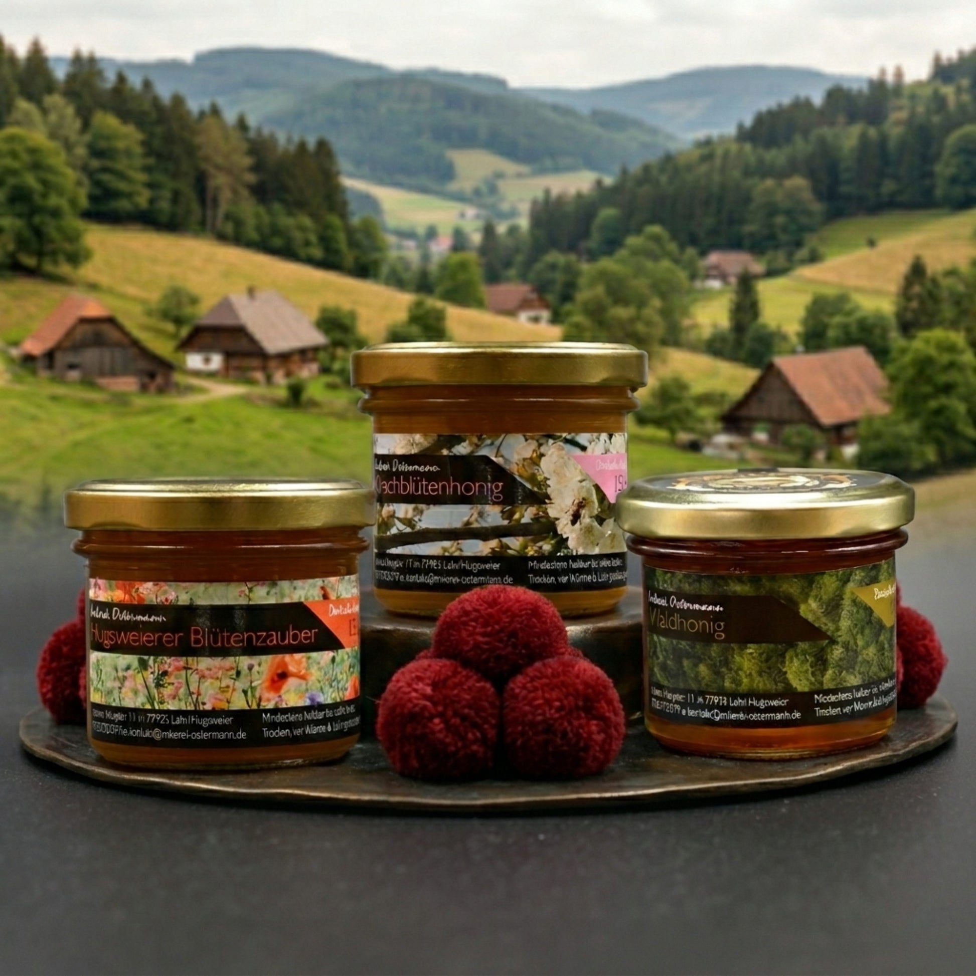Drei Gläser Honig der Imkerei Ostermann auf einem dunklen Holzteller vor einer malerischen Schwarzwaldkulisse. Das Set "Badisches Versucherle" enthält Hugswierer Blütenzauber, Kirschblütenhonig und Waldhonig, dekoriert mit roten Bollenhut-Pompons.