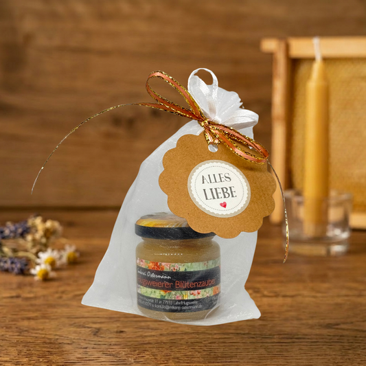 Ein kleines Glas Honig der Imkerei Ostermann (Sorte "Blütenzauber") auf einem rustikalen Holztisch ist in einem weißen, durchsichtigen Organzasäckchen verpackt.