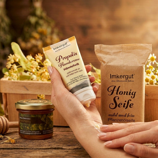 Handpflege-Geschenkset Süße Pflege der Imkerei Ostermann: Gepflegte Hände tragen die Propolis Handcreme auf, daneben stehen die Honigseife in Papierverpackung und ein Glas Waldhonig auf einem Holztisch, dekoriert mit Kamillenblüten.